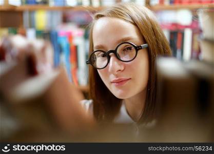 People: young girl, student, wearing eyeglasses, chooses a book in a library or bookstore, smiling.. Girl and books