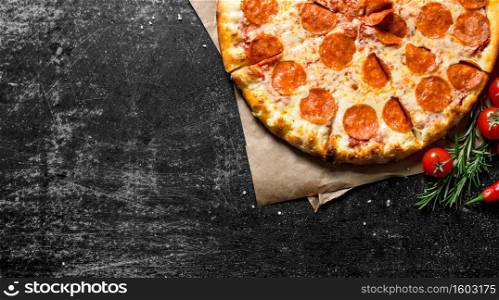 Pepperoni pizza on paper with tomatoes and rosemary. On dark rustic background. Pepperoni pizza on paper with tomatoes and rosemary.