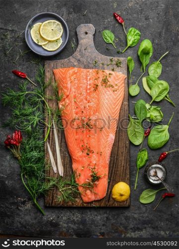 Perfect salmon fillet on rustic cutting board with fresh ingredients for tasty cooking on dark background, top view. Healthy or diet food concept.
