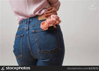 Perfect woman's bottom in a blue jeans and fresh roses living coral color in a back pocket on a light gray background, place for text. Concept of Woman's or Mother's Day.. Attractive woman's butt in a blue jeans and fresh roses living coral color in a back pocket on a light gray background, place for text. Congratulation card.