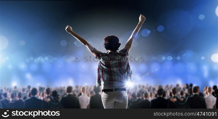 Performer on stage. Back view of girl with hands up standing in stage lights