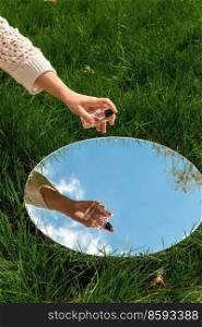 perfumery and nature concept - hand with bottle of perfume and sky reflection in round mirror on summer field. hand with perfume and sky reflection in mirror