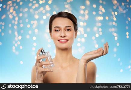 perfumery, beauty and luxury concept - happy smiling young woman with perfume over holidays lights on blue background. happy woman with perfume