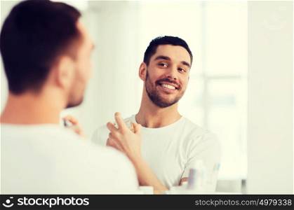 perfumery, beauty and people concept - happy smiling young man with perfume looking to mirror using scent at bathroom. man with perfume looking to mirror at bathroom