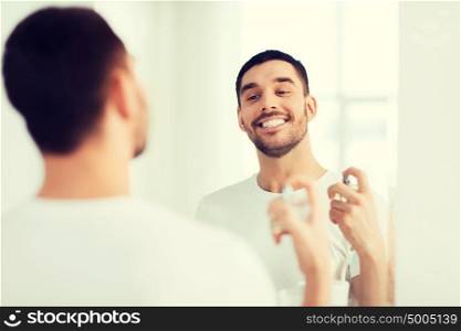 perfumery, beauty and people concept - happy smiling young man with perfume looking to mirror using scent at bathroom. man with perfume looking to mirror at bathroom