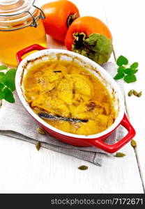 Persimmon baked with honey, cardamom and vanilla in a roasting pan on a towel, mint and orange fruit on light wooden board