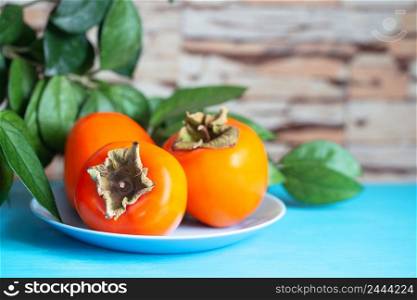 Persimmon on a blue plate against a background of green foliage and a stone wall. Selective focus. Persimmon on blue plate against background of green foliage and stone wall