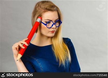 Person confused thinking seeks a solution. Pensive thoughtful student girl or business woman female teacher wearing nerdy glasses coming up with an idea, holding big red pencil. Studio shot on grey.. Woman confused thinking, big pencil in hand