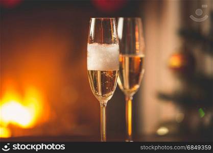 Person filling two glasses with champagne. Burning fireplace and decorated Christmas tree