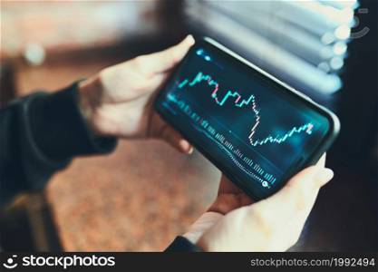 Person investing trading on stock cryptocurrency market using investing application on smartphone. Stock market investment in hand. Trader holding mobile phone looking at candle chart