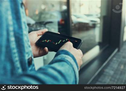 Person investing trading on stock cryptocurrency market using investing application on smartphone. Stock market investment in hand. Trader holding mobile phone looking at candle chart. Man investing trading on stock cryptocurrency market using investing application on smartphone. Stock market investment in hand. Trader holding mobile phone looking at candle chart