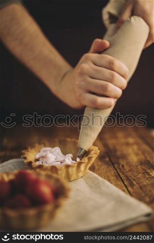 person s hand filling tart with pink whipped cream from icing bag wooden table