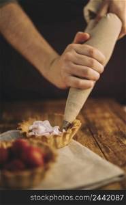 person s hand filling tart with pink whipped cream from icing bag wooden table