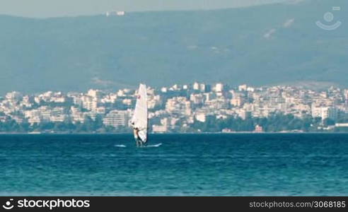 Person sailboarding offshore on a calm ocean with a town visible on the coastline in the distance behind