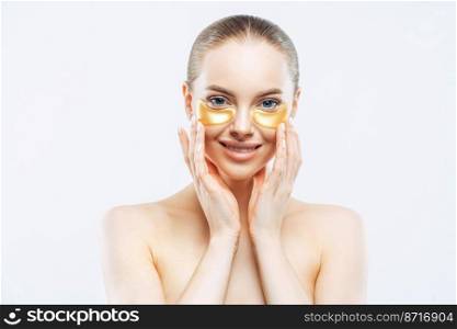 Personal hygiene, beauty, wellness concept. Tender smiling European woman touches face, applies under eye patches, collagen mask for fresh facial skin, stands topless against white background