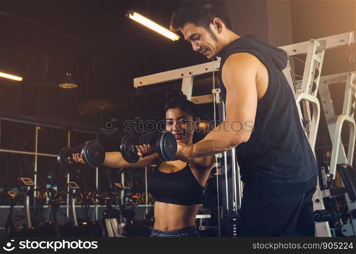 Personal training man helping women's arms to pull up dumbbell for exercise at indoor gym.