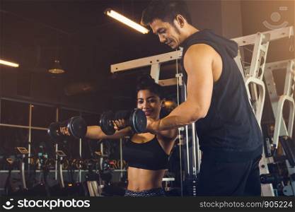 Personal training man helping women's arms to pull up dumbbell for exercise at indoor gym.