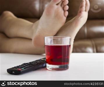 Persons feet up on coffee table while watching TV with remote control and drink on table.