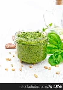 Pesto sauce in a glass jar, basil, pine nuts, garlic and olive oil in a carafe on background of white wooden board
