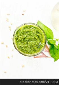Pesto sauce in a glass jar, basil, pine nuts, garlic and olive oil in a carafe on the background of a light wooden board on top