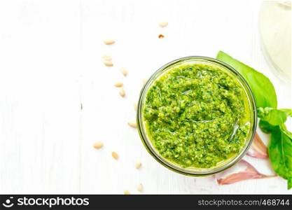 Pesto sauce in a glass jar, basil, pine nuts, garlic and olive oil in a carafe on wooden board background from above