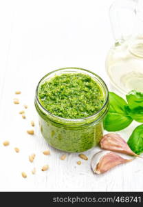 Pesto sauce in a glass jar, pine nuts, garlic, green basil and olive oil in a carafe on the background of light wooden board