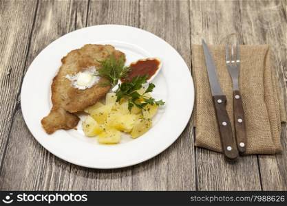 Petite Wiener schnitzel with boiled potatoes and ketchup. Served on a white porcelain plate with fork and knife on a wooden background.. Petite Wiener schnitzel with boiled potatoes and ketchup. Served on a white porcelain plate with fork and knife on a wooden background