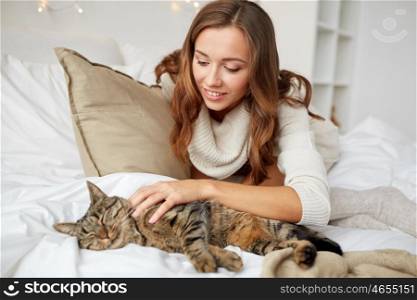 pets, comfort, christmas, winter and people concept - happy young woman with cat lying in bed at home