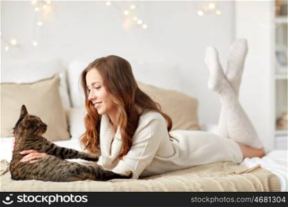 pets, comfort, christmas, winter and people concept - happy young woman with cat lying in bed at home