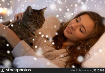 pets, comfort, rest and people concept - happy young woman with cat lying in bed at home at night over snow. happy young woman with cat lying in bed at home