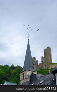 Pfarrkirche church and Lowenburg castle with flying birds in the background at the picturesque village of Monreal in the Eifel region, Germany