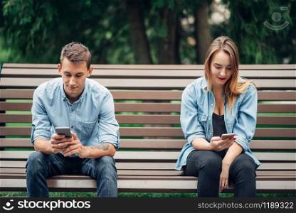 Phone addicted couple on the bench in park. Man and woman using their smartphones, addiction