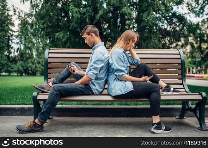Phone addicted people, couple on the bench. Man and woman using their smartphones in summer park, addiction