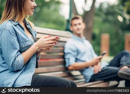 Phone addiction, smiling couple on the bench in park. Man and woman using their smartphones, social addicted people