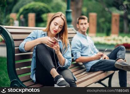 Phone addiction, smiling couple on the bench in park. Man and woman using their smartphones, social addicted people. Phone addiction, smiling couple on bench in park