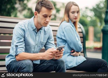 Phone addiction, young couple on the bench in park. Man and woman using their smartphones, addicted people. Phone addiction, young couple on the bench in park