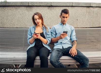 Phone addiction, young couple on the bench in park. Man and woman using their smartphones, addicted people. Phone addiction, young couple on the bench in park
