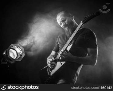 Photo of a bearded man playing electric guitar on stage in front of spotlights.