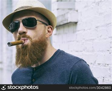 Photo of a bearded man with hat and sunglasses standing outside smoking a cigar.. Bearded man with sunglasses smoking a cigar
