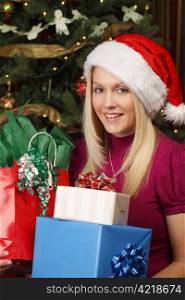 Photo of a beautiful blond female sitting in front of a Christmas tree holding presents.