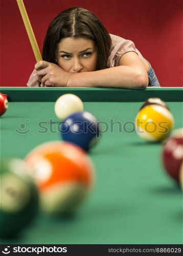 Photo of a beautiful brunette at the edge of a billiards table holding a pool cue and wondering about her next shot.