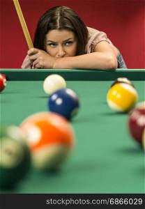Photo of a beautiful brunette at the edge of a billiards table holding a pool cue and wondering about her next shot.
