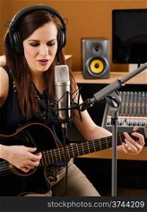 Photo of a beautiful brunette in a recording studio playing an acoustic guitar and singing into a large diaphragm microphone.