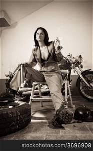 Photo of a beautiful female mechanic wearing overalls, leather bra, and sitting in front of a motorcycle smoking and holding a tool.