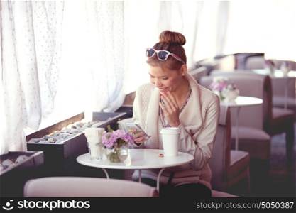 Photo of a beautiful happy young woman using wireless internet, surfing the net via smartphone and drinking coffee in a cafe in the morning.