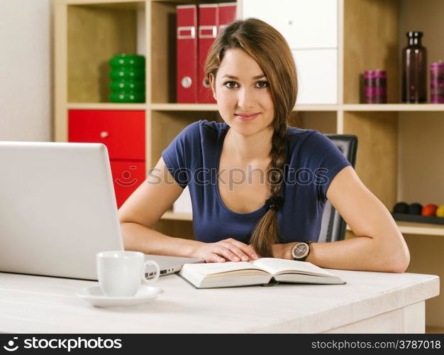 Photo of a beautiful smiling woman using a laptop and drinking coffee at home or at her office.