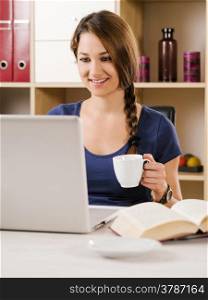 Photo of a beautiful smiling woman using a laptop and drinking coffee at home or at her office.