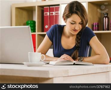 Photo of a beautiful woman using a laptop, drinking coffee, and reading a book at home or an office.