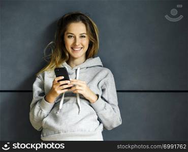 Photo of a beautiful young blond woman leaning up against a wall, smiling while using her cellphone.
