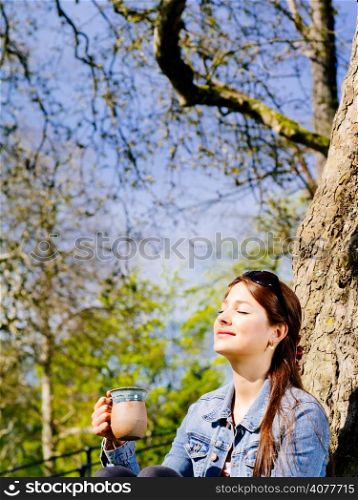 Photo of a beautiful young woman drinking coffee or tea while sitting outdoors enjoying the warmth of the sun on her face.
